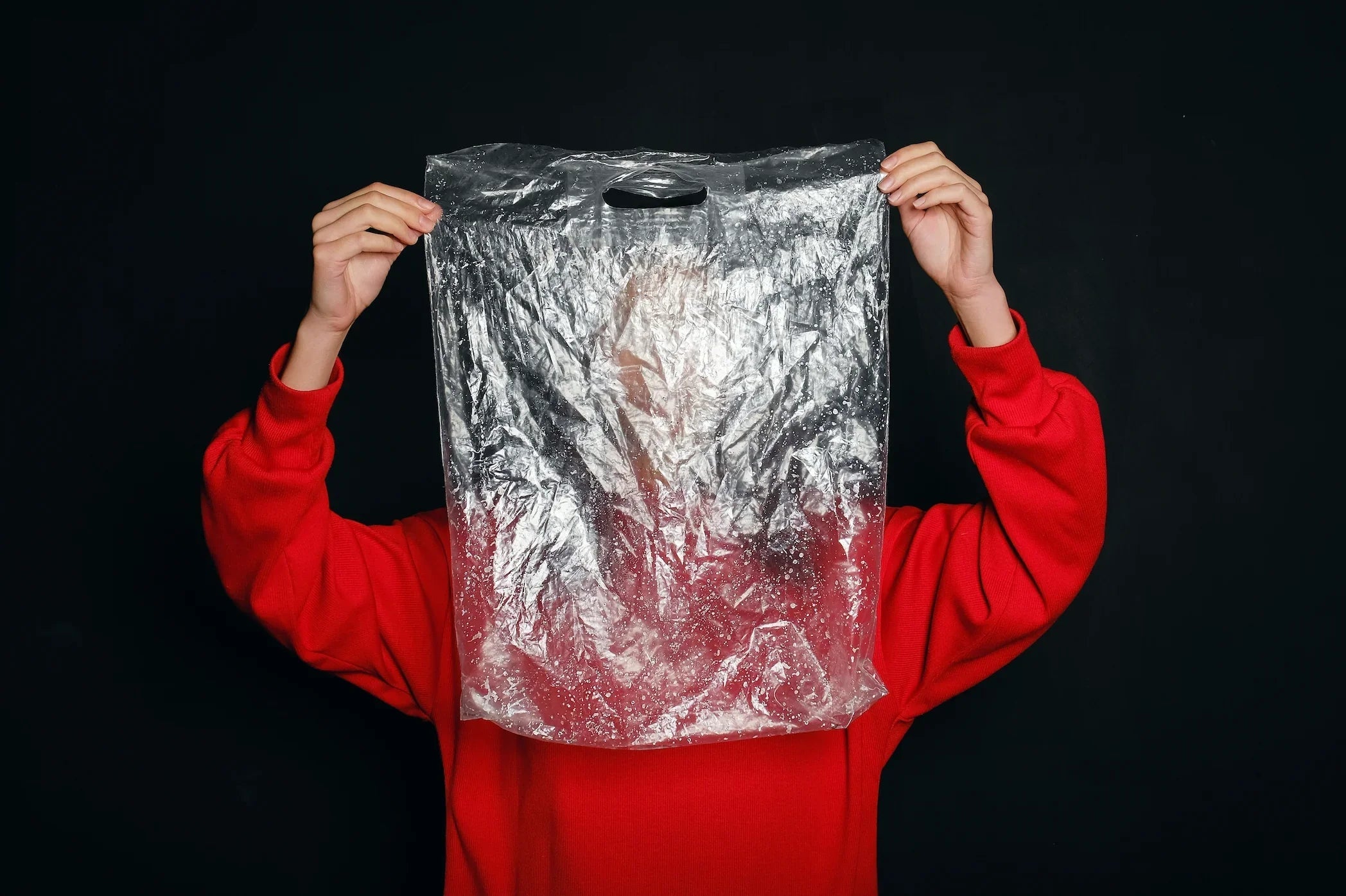 Person in red sweater holding large clear plastic bag against black background
