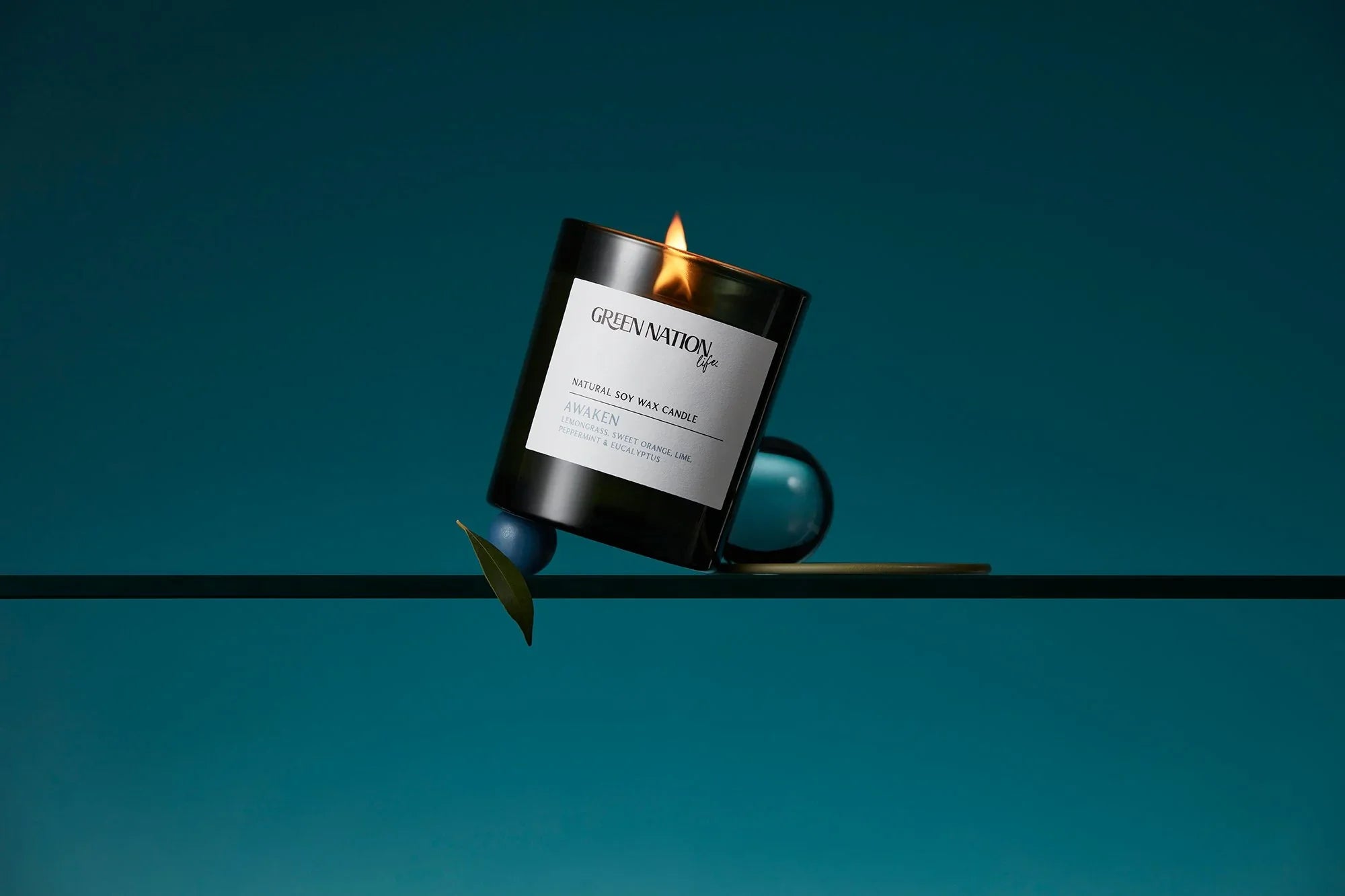 Green Nation Life black soy wax candle labeled Awaken, burning on a glass shelf with blue background