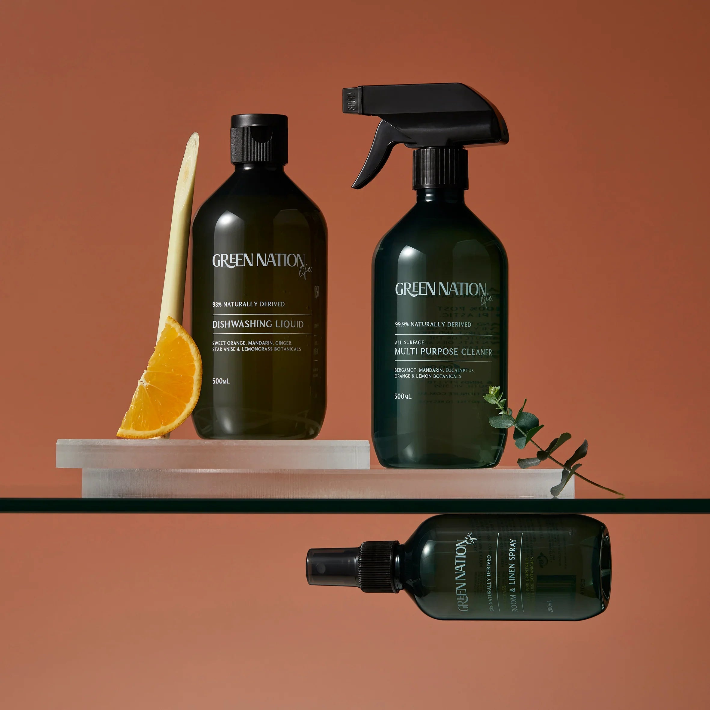 Green Nation dishwashing liquid, multi purpose cleaner, and room spray bottles with orange and lemongrass on a glass shelf, against a brown background.